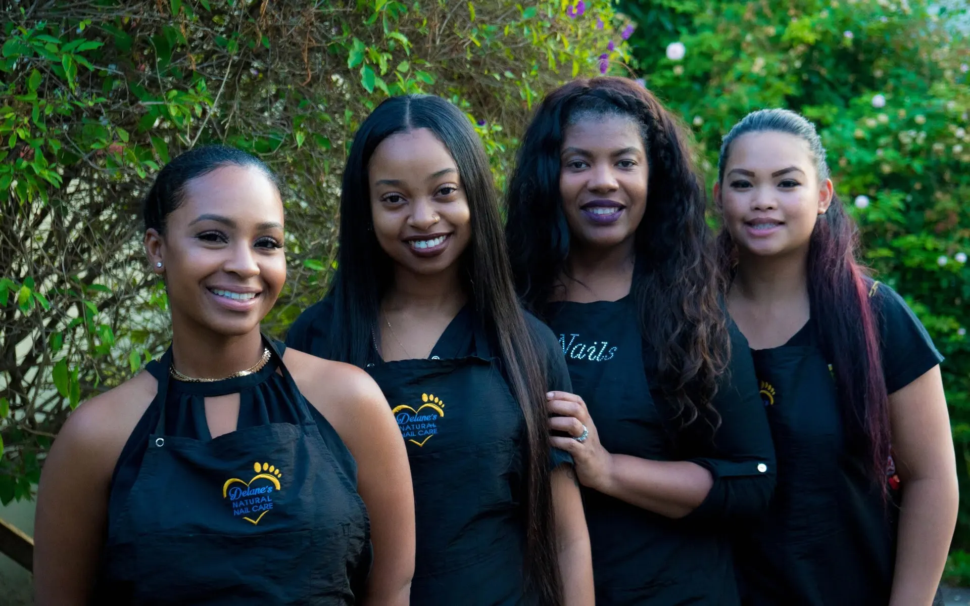 Four smiling women in black aprons and tops, three of them embroidered with the Delane's Natural Nail Care logo, standing together outdoors in front of green foliage