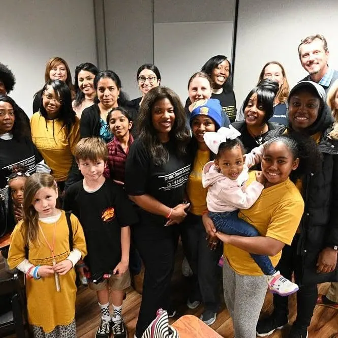 A large group of women, men and children posing together indoors; several wear yellow or black DNNC Steps to Success shirts
