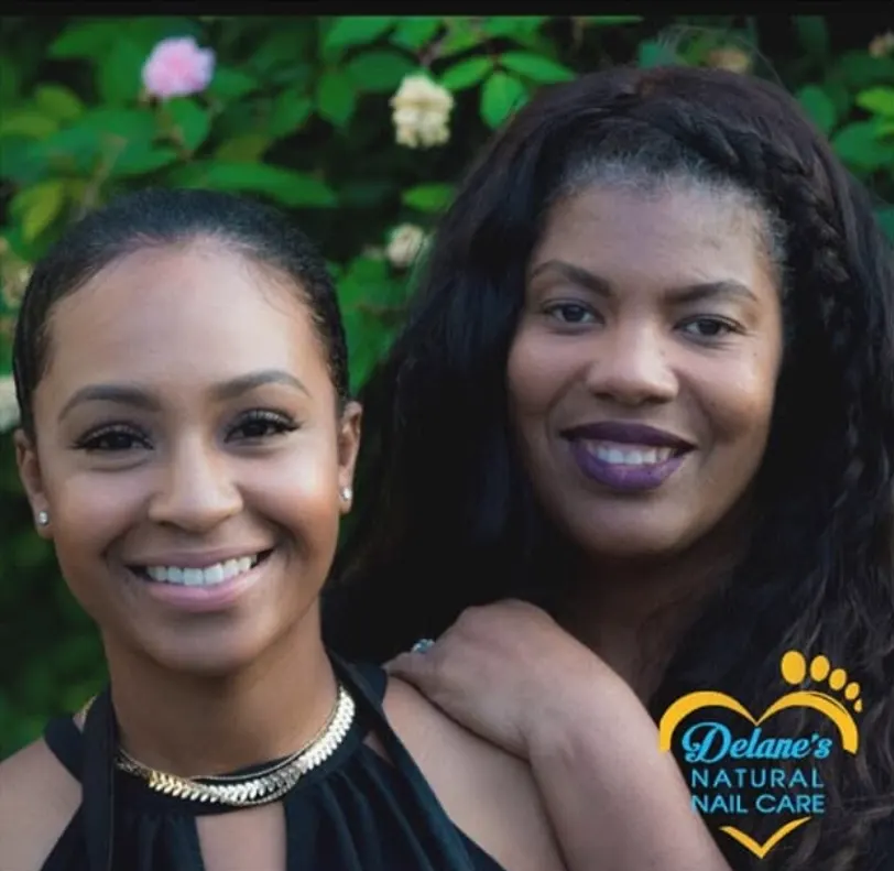 Two women smiling close together in a garden, with the Delane's Natural Nail Care logo in the lower corner