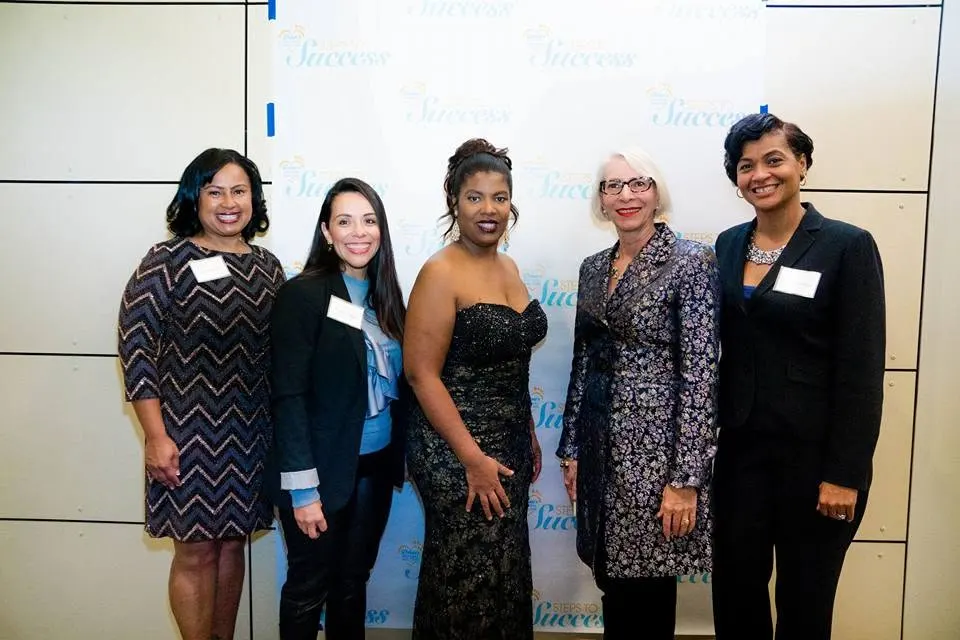 Five women in professional and evening attire posing in front of a Steps to Success step-and-repeat backdrop