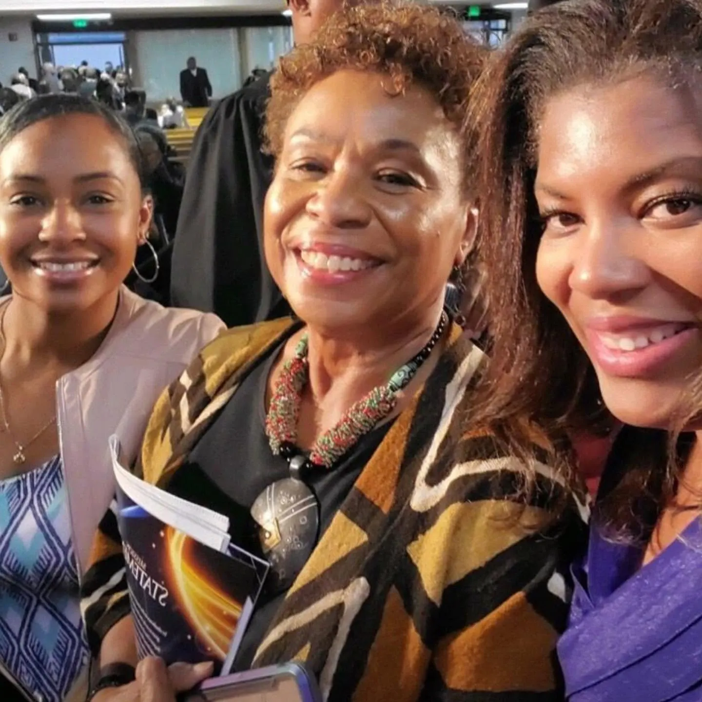 Three smiling women taking a close selfie in a crowded auditorium
