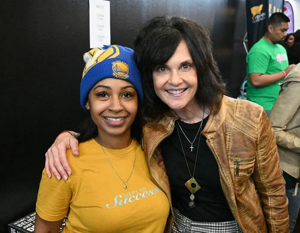 A woman in a Warriors beanie and a yellow Steps to Success shirt posing with a woman in a tan leather jacket at a community event