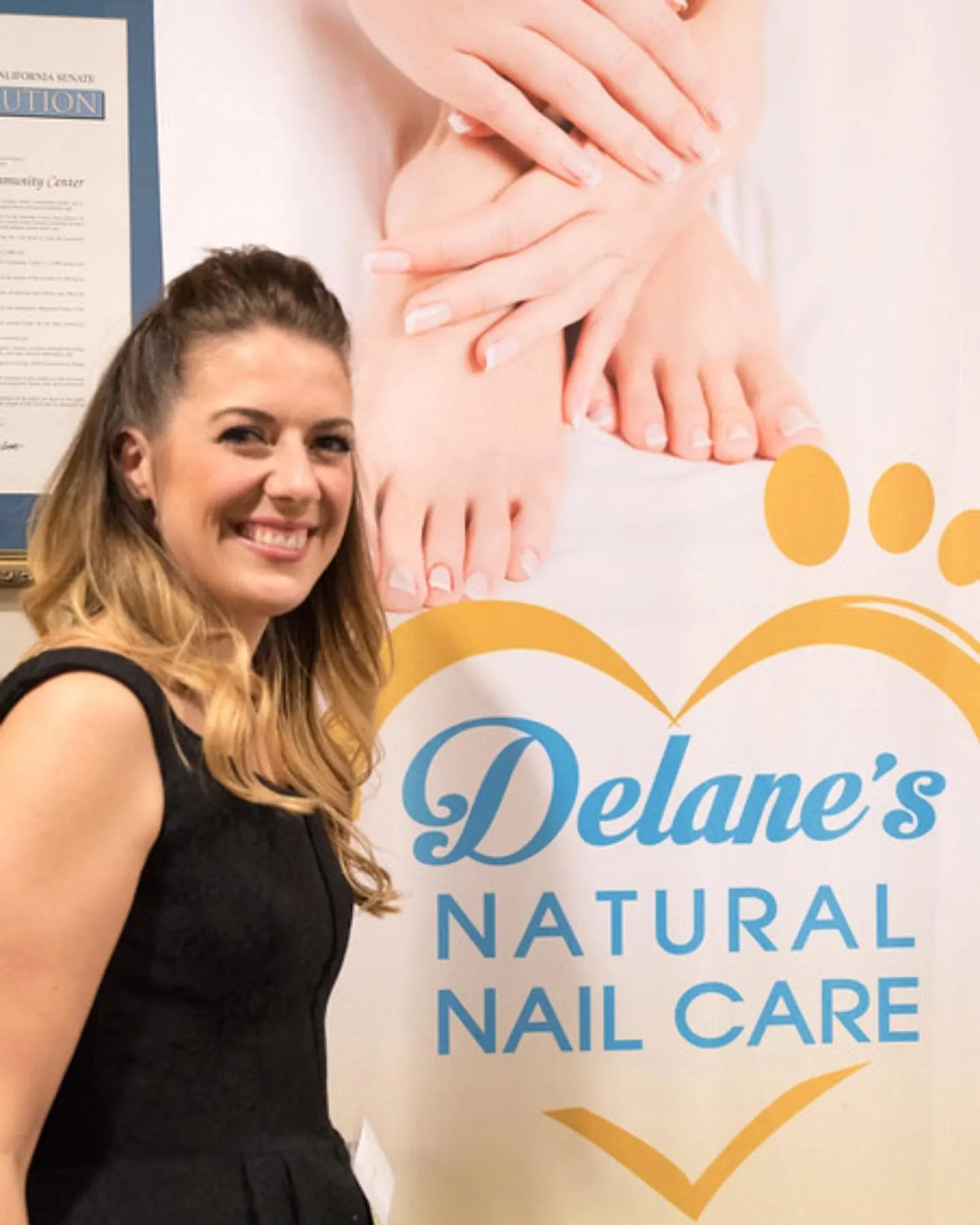A smiling woman in a black dress standing beside a Delane's Natural Nail Care banner showing hands and feet