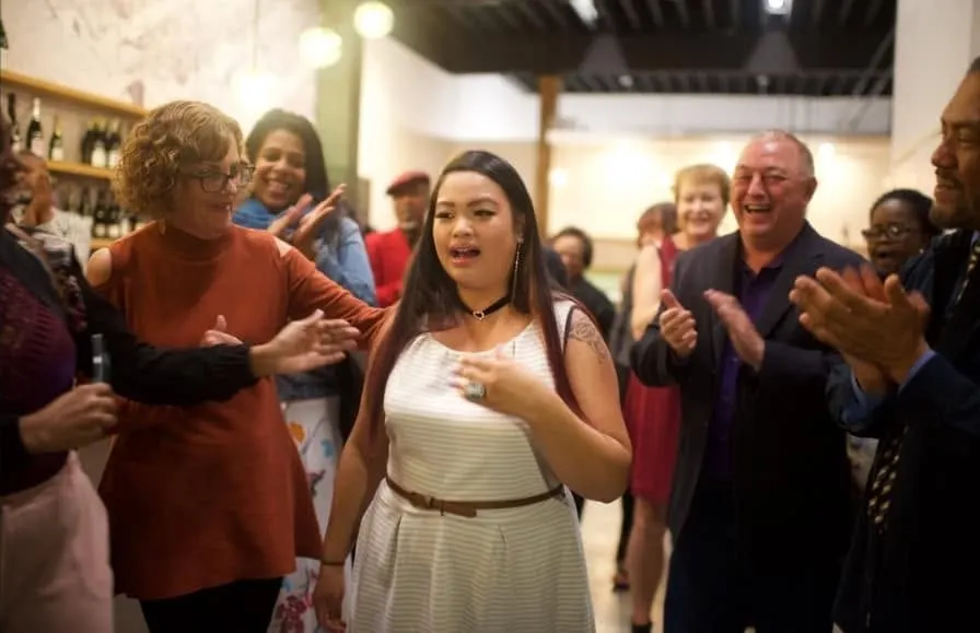 A woman in a white dress smiling and pressing her hands together while guests around her applaud