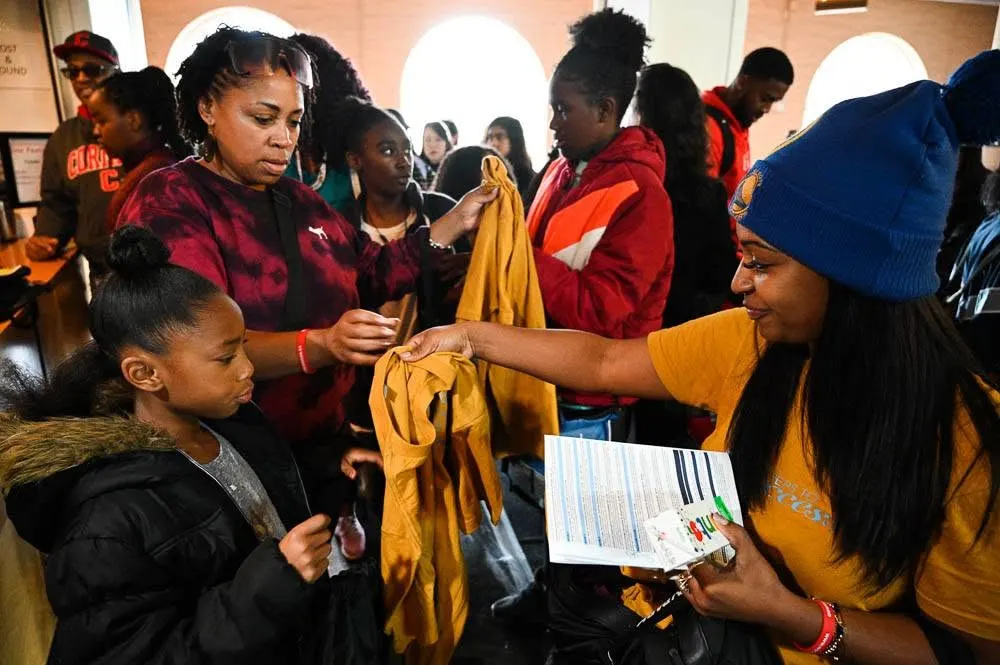 Women and children gathered indoors looking through donated clothing, with one woman in a blue knit cap holding a yellow garment