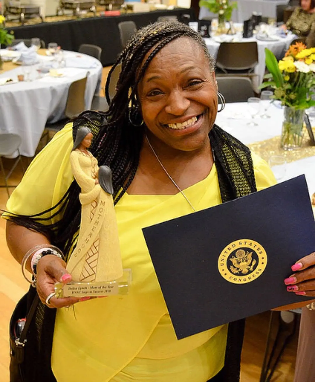 A smiling woman in yellow holding a statuette and a navy folder with a U.S. Congress seal at a banquet